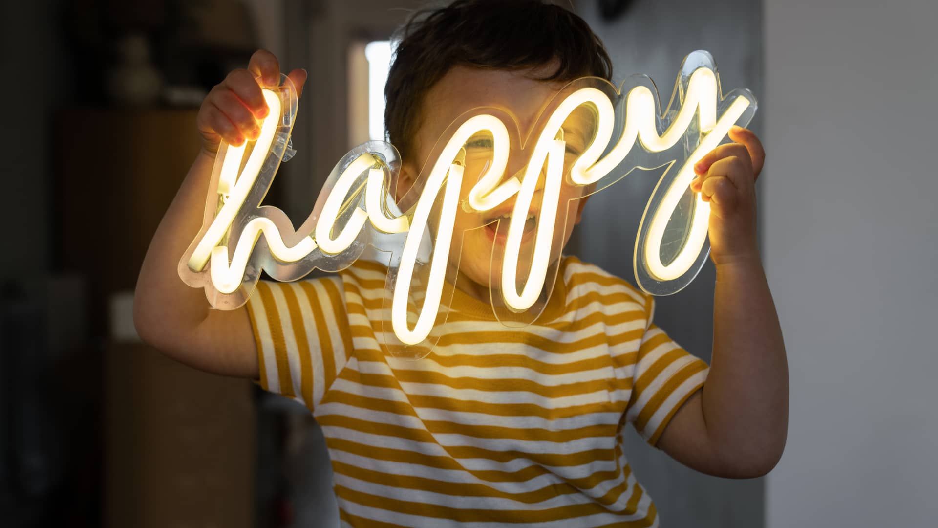 Niño sonriendo sosteniendo palabra luminosa LED happy, un tipo de luz que permite ahorrar sobre otras Niño sonriendo sosteniendo palabra luminosa LED happy, un tipo de luz que permite ahorrar sobre otras